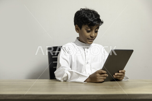 الجلوس على المكتب والاستمتاع باستخدام الجهاز اللوحي, متابعة البرامج التعليمية والترفيهية بإيماءات السرور، بورتريه لولد عربي خليجي سعودي مبتسم يرتدي الثوب التقليدي ويحمل بيده التابليت، استخدام الأجهزة المحمولة في التعلم والترفيه، خلفية بيضاء
