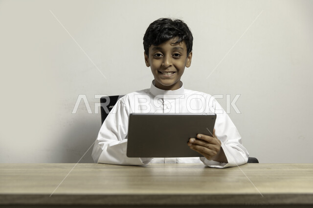 الجلوس على الطاولة والنظر الى الكاميرا, متابعة البرامج التعليمية والترفيهية بإيماءات السرور، بورتريه لولد عربي خليجي سعودي مبتسم يرتدي الثوب التقليدي ويحمل بيده جهاز التابليت، استخدام الأجهزة المحمولة في التعلم والترفيه، خلفية بيضاء