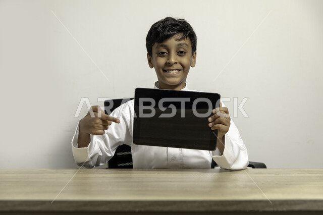 الإشارة الى الجهاز اللوحي بشاشة سوداء فارغة، بورتريه لصبي عربي خليجي سعودي مبتسم يرتدي الثوب التقليدي يجلس على الطاولة وينظر الى الكاميرا و يحمل بيده التابلت، استعمال التقنيات الحديثة للتسويق الرقمي الالكتروني، عرض تطبيقات وخدمات الانترنت, خلفية بيضاء