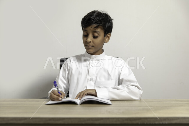 الاجتهاد والاستعداد للإختبارات المدرسية بإيماءات التركيز, بورتريه لصبي عربي خليجي سعودي يرتدي الثوب السعودي يجلس على الطاولة ويقوم بحل الواجبات المنزلية, مفهوم التعلم و التثقف الذاتي, تنمية  وتطوير مهارات الكتابة, خلفية بيضاء