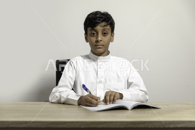 The concept of learning and self-cultivation, a portrait of a Saudi Gulf Arab boy wearing the Saudi dress, sitting at the table, doing homework and looking at the camera, diligence and preparing for school tests, developing writing skills, white background