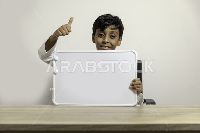 التسويق على لوح إعلاني فارغ بإيماءات الإعجاب والرضا, بورتريه لولد عربي سعودي خليجي مبتسم يرتدي الثوب التقليدي يجلس على طاولته ويحمل بيده لوح أبيض فارغ, الإعلان عن بدء عام دراسي جديد في مدارس المملكة العربية السعودية, خلفية بيضاء