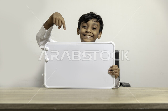 الترويج على لوح إعلاني فارغ, النظر الى الكاميرا بإيماءات السعادة, بورتريه لولد عربي سعودي خليجي مبتسم يرتدي الثوب التقليدي يجلس على طاولته ويشير بيده الى لوح أبيض فارغ, الإعلان عن بدء عام دراسي جديد في مدارس المملكة العربية السعودية, خلفية بيضاء
