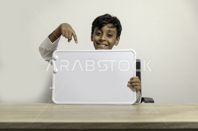 الترويج على لوح إعلاني فارغ, النظر الى الكاميرا بإيماءات السعادة, بورتريه لولد عربي سعودي خليجي مبتسم يرتدي الثوب التقليدي يجلس على طاولته ويشير بيده الى لوح أبيض فارغ, الإعلان عن بدء عام دراسي جديد في مدارس المملكة العربية السعودية, خلفية بيضاء