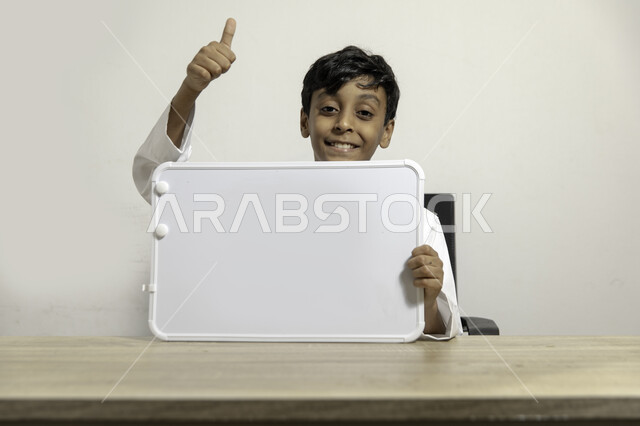 التسويق على لوح إعلاني فارغ بإيماءات الإعجاب والرضا, بورتريه لولد عربي سعودي خليجي مبتسم يرتدي الثوب التقليدي يجلس على طاولته ويحمل بيده لوح أبيض فارغ, الإعلان عن بدء عام دراسي جديد في مدارس المملكة العربية السعودية, خلفية بيضاء
