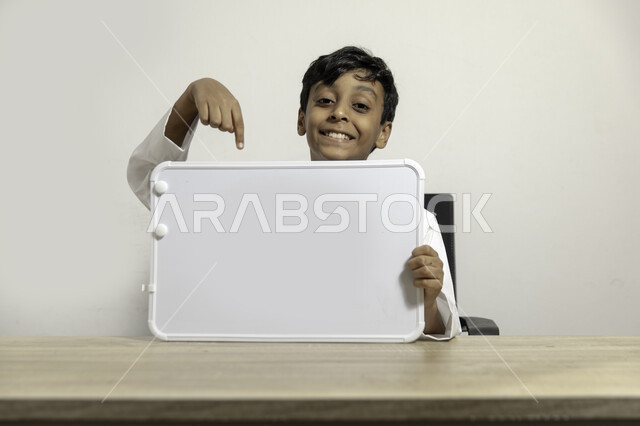 النظر الى الكاميرا بإيماءات السعادة, الترويج على لوح إعلاني فارغ, بورتريه لولد عربي سعودي خليجي مبتسم يرتدي الثوب التقليدي يجلس على طاولته ويشير بيده الى لوح أبيض فارغ, الإعلان عن بدء عام دراسي جديد في مدارس المملكة العربية السعودية, خلفية بيضاء