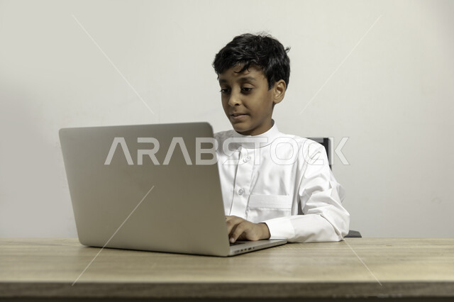 متابعة الدروس والشروحات عن بعد, بورتريه لصبي عربي خليجي سعودي يرتدي الثوب التقليدي يجلس على الطاولة ويستخدم اللابتوب, استعمال الأجهزة التقنية في التعلم, التسلية والترفيه بقضاء الوقت بتصفح مواقع التواصل الاجتماعي على الحاسوب المحمول, خلفية بيضاء