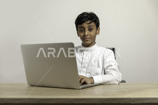 Shopping and payment through mobile applications, modern and advanced technologies, close-up portrait of a Saudi Arabian Gulf child wearing traditional dress sitting on a chair holding a laptop and a bank credit card, paying bills with electronic cards, beige background