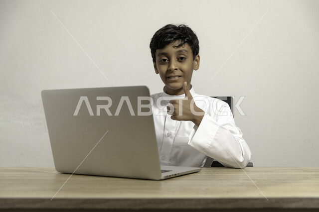رفع أصبع الإبهام بإيماءات القبول والإعجاب, متابعة الدروس والشروحات عن بعد, بورتريه لصبي عربي خليجي سعودي مبتسم يرتدي الثوب التقليدي يجلس على الطاولة ويستخدم اللابتوب, استعمال الأجهزة التقنية في التعلم, التسلية والترفيه بقضاء الوقت بتصفح التطبيقات , خلفية بيضاء