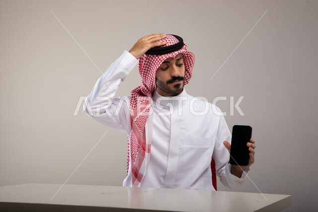 انجاز مهام العمل عن بعد, بورتريه لرجل عربي خليجي سعودي يرتدي الثوب التقليدي والشماغ يمسك الجوال ويقوم برفع يديه على رأسه بإيماءات القلق, عرض تطبيقات واستخدامات الانترنت في مجال العمل المكتبي, خلفيه رماديه