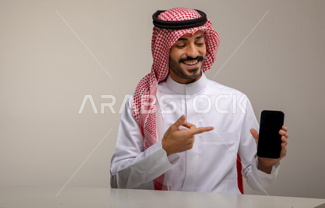 الجلوس في المكتب ومتابعه أخبار وسائل السوشال ميديا باستخدام الجوال, انجاز مهام العمل عبر الانترنت بإيماءات السرور, بورتريه لرجل عربي خليجي سعودي مبتسم يرتدي الزي التقليدي يشير بإصبعه السبابة إلى الهاتف المحمول بشاشه سوداء فارغه, خلفيه رماديه
