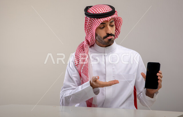 اكتشاف مشاكل وأعطال الهاتف المحمول, بورتريه لشاب عربي خليجي سعودي يرتدي الثوب التقليدي والشماغ يجلس في مكتبه يمسك جوال بشاشه سوداء فارغه ويشير إليه, استخدام التقنيات الحديية المتطورة في انجاز الأعمال, خلفيه رماديه