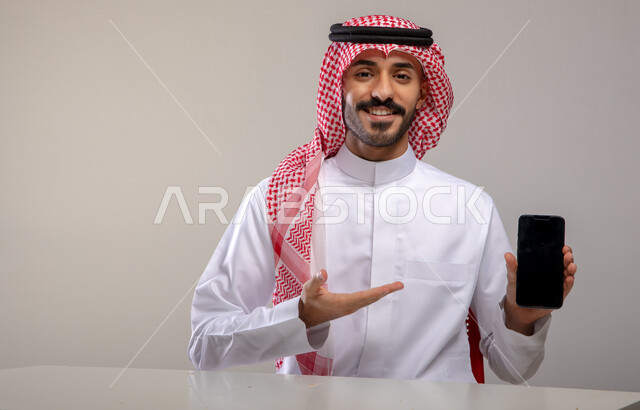 عرض تطبيقات وخدمات الهاتف المحمول, بورتريه لشاب عربي خليجي سعودي مبتسم يرتدي الثوب التقليدي والشماغ يحمل بيده جوال بشاشه سوداء فارغة ويشير إليه, النظر إلى الكاميرا بإيماءات السرور, استخدام التكنولوجيا الحديثة للتسوق الالكتروني الذكي عبر الانترنت, خلفيه رماديه