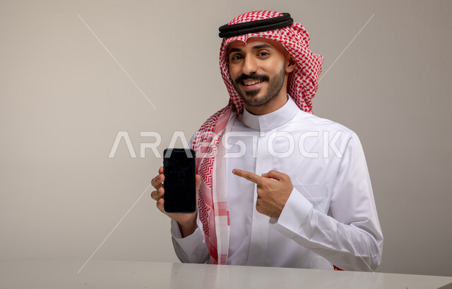 انجاز مهام العمل عبر الانترنت, بورتريه لرجل عربي خليجي سعودي مبتسم يرتدي الزي التقليدي يشير بإصبعه السبابة إلى الهاتف المحمول بشاشه سوداء فارغه, متابعه أخبار وسائل السوشال ميديا باستخدام الجوال, الجلوس في المكتب والنظر إلى الكاميرا, خلفيه رماديه