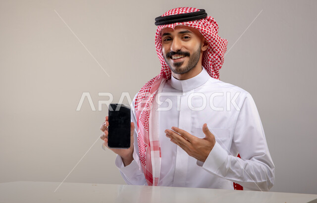 استخدام التكنولوجيا الحديثة للتسوق الالكتروني عبر الانترنت, عرض تطبيقات وخدمات الهاتف المحمول, بورتريه لشاب عربي خليجي سعودي مبتسم يرتدي الثوب التقليدي والشماغ يحمل بيده جوال بشاشه سوداء فارغة ويشير إليه, النظر إلى الكاميرا بإيماءات السرور, خلفيه رماديه