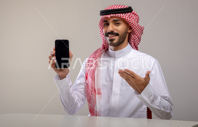 عرض تطبيقات وخدمات الهاتف المحمول, بورتريه لشاب عربي خليجي سعودي مبتسم يرتدي الثوب التقليدي والشماغ يحمل بيده جوال بشاشه سوداء فارغة ويشير إليه, النظر إلى الكاميرا بإيماءات السرور, استخدام التكنولوجيا الحديثة للتسوق الالكتروني عبر الانترنت, خلفيه رماديه