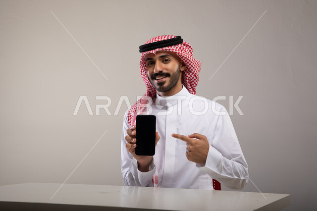 الجلوس في المكتب والنظر إلى الكاميرا, انجاز مهام العمل عبر الانترنت, بورتريه لرجل عربي خليجي سعودي مبتسم يرتدي الزي التقليدي يشير بإصبعه السبابة إلى الهاتف المحمول بشاشه سوداء فارغه, متابعه أخبار وسائل السوشال ميديا باستخدام الجوال, خلفيه رماديه