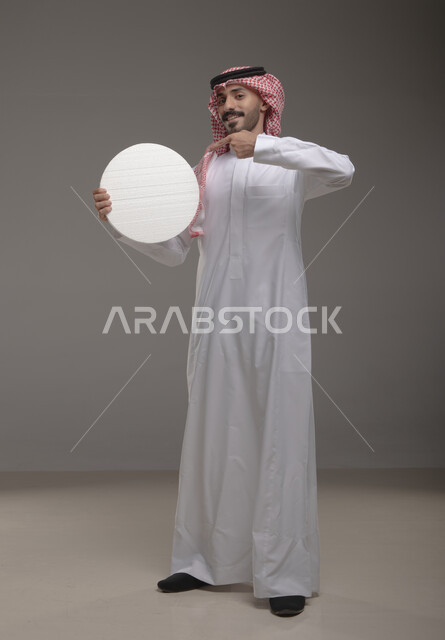 الوقوف باستقامه وثقه ، موك اب دائري أبيض ،  بورتريه لرجل عربي سعودي خليجي يرتدي الشماغ والثوب التقليدي مبتسم يحمل بيده لوحه بيضاء مدوره، لوح إعلاني فارغ،  صورة الجسم بالطول الكامل، النظر الى الكاميرا بإيماءات الإعجاب والسرور، خلفية رماديه