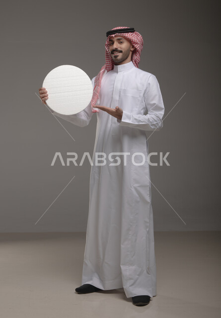 لوح إعلاني فارغ، موك اب دائري أبيض ،  بورتريه لرجل عربي سعودي خليجي يرتدي الشماغ والثوب التقليدي مبتسم يحمل بيده لوحه بيضاء مدوره،  الوقوف باستقامه وثقه، صورة الجسم بالطول الكامل، النظر الى الكاميرا بإيماءات الإعجاب والسرور، خلفية رماديه