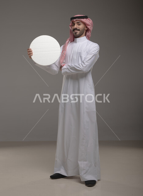 موك اب دائري أبيض ،  بورتريه لرجل عربي سعودي خليجي يرتدي الشماغ والثوب التقليدي مبتسم يحمل بيده لوحه بيضاء مدوره، لوح إعلاني فارغ، الوقوف باستقامه وثقه، صورة الجسم بالطول الكامل، النظر الى الكاميرا بإيماءات الإعجاب والسرور، خلفية رماديه