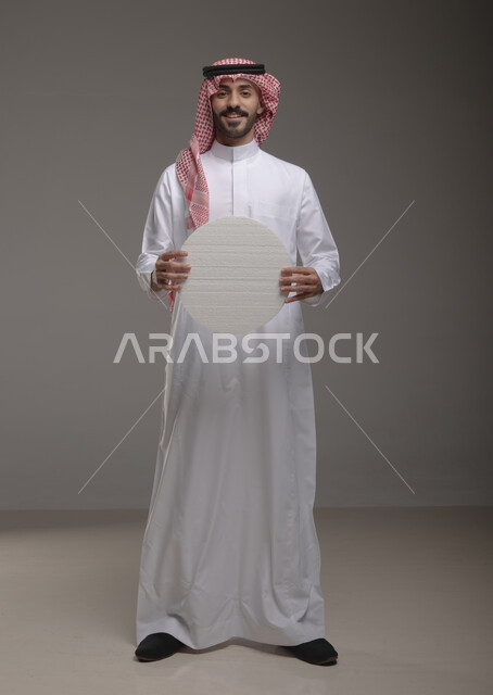 الوقوف باستقامة وثقة ، موك اب دائري أبيض ،  بورتريه لرجل عربي سعودي خليجي يرتدي الشماغ والثوب التقليدي مبتسم يحمل بيده لوحه بيضاء مدوره، لوح إعلاني فارغ، صورة الجسم بالطول الكامل، النظر الى الكاميرا بإيماءات الإعجاب والسرور، خلفية رماديه