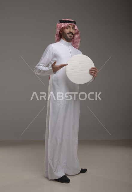 موك اب دائري أبيض ،  بورتريه لرجل عربي سعودي خليجي يرتدي الشماغ والثوب التقليدي مبتسم يحمل بيده لوحه بيضاء مدوره، لوح إعلاني فارغ، الوقوف باستقامه وثقه، صورة الجسم بالطول الكامل، النظر الى الكاميرا بإيماءات الإعجاب والسرور، خلفية رماديه