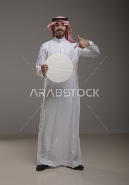 إيماءات الإعجاب والسرور والنظر إلى الكاميرا ، موك اب دائري أبيض ،  بورتريه لرجل عربي سعودي خليجي يرتدي الشماغ والثوب التقليدي مبتسم يحمل بيده لوحه بيضاء مدوره، لوح إعلاني فارغ، الوقوف باستقامه وثقه، صورة الجسم بالطول الكامل، خلفية رماديه