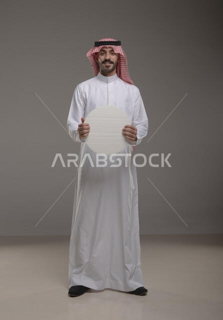 الوقوف بثقه واستقامه ، موك اب دائري أبيض ،  بورتريه لرجل عربي سعودي خليجي يرتدي الشماغ والثوب التقليدي مبتسم يحمل بيده لوحه بيضاء مدوره، لوح إعلاني فارغ،  صورة الجسم بالطول الكامل، النظر الى الكاميرا بإيماءات الإعجاب والسرور، خلفية رماديه