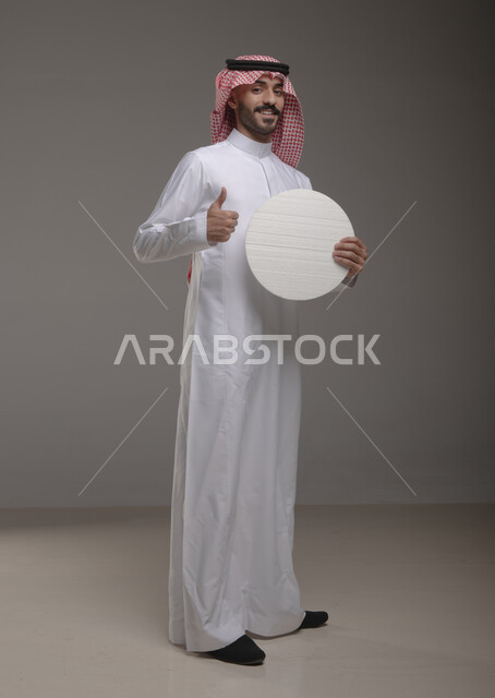 السرور والإعجاب إيماءات النظر إلى الكاميرا ، موك اب دائري أبيض ،  بورتريه لرجل عربي سعودي خليجي يرتدي الشماغ والثوب التقليدي مبتسم يحمل بيده لوحه بيضاء مدوره، لوح إعلاني فارغ، الوقوف باستقامه وثقه، صورة الجسم بالطول الكامل،  خلفية رماديه