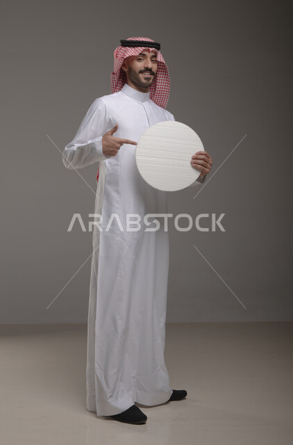 الاستقامه والوقوف بثقه ، موك اب دائري أبيض ،  بورتريه لرجل عربي سعودي خليجي يرتدي الشماغ والثوب التقليدي مبتسم يحمل بيده لوحه بيضاء مدوره، لوح إعلاني فارغ، صورة الجسم بالطول الكامل، النظر الى الكاميرا بإيماءات الإعجاب والسرور، خلفية رماديه