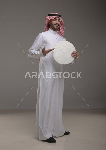 موك اب دائري أبيض ،  بورتريه لرجل عربي سعودي خليجي يرتدي الشماغ والثوب التقليدي مبتسم يحمل بيده لوحه بيضاء مدوره، لوح إعلاني فارغ، الوقوف باستقامه وثقه، صورة الجسم بالطول الكامل، النظر الى الكاميرا بإيماءات الإعجاب والسرور، خلفية رماديه