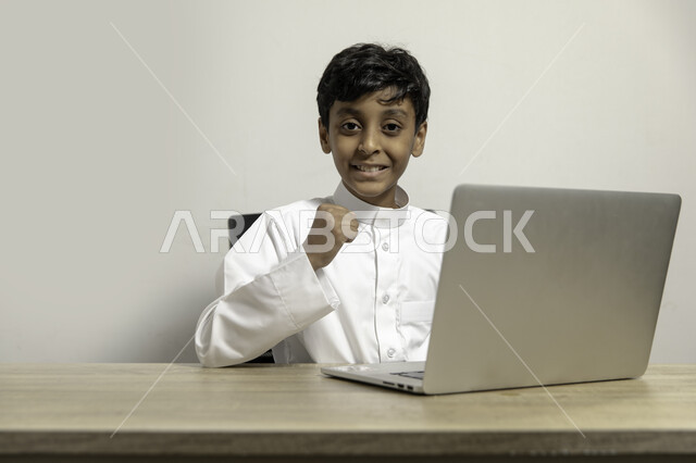 استعمال الأجهزة التقنية في دراسة المقررات المدرسية عن بعد, متابعة نتائج الامتحان عبر جهاز الحاسوب المحمول, بورتريه لصبي عربي خليجي سعودي يرتدي الثوب التقليدي يجلس على الطاولة وينظر الى الكاميرا ويستخدم اللابتوب, إيماءات الفرح بالنجاح و التفوق, خلفية بيضاء