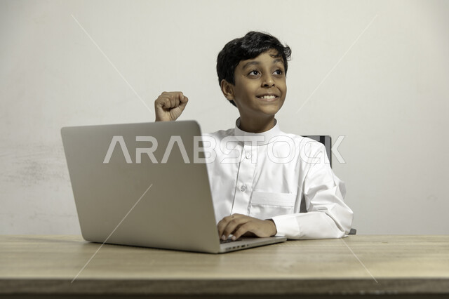 إيماءات النظر الى الأعلى و الفرح بالنجاح و التفوق, متابعة نتائج الامتحان عبر الحاسوب المحمول, دراسة المقررات المدرسية عن بعد, بورتريه لصبي عربي خليجي سعودي يرتدي الثوب التقليدي يجلس على الطاولة ويستخدم اللابتوب, استعمال الأجهزة التقنية في التعلم, خلفية بيضاء
