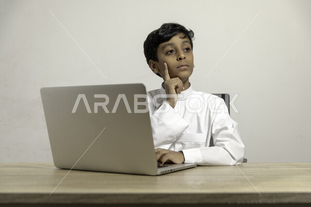 إيماءات التفكير بالمستقبل والحلول, متابعة الدروس والشروحات عن بعد, بورتريه لصبي عربي خليجي سعودي يرتدي الثوب التقليدي يجلس على الطاولة ويستخدم اللابتوب, استعمال الأجهزة التقنية في التعلم, خلفية بيضاء