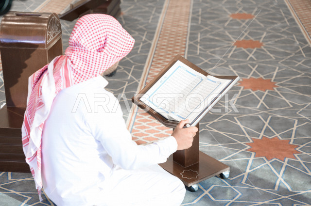 رجل سعودي مسلم يجلس داخل المسجد و يقرأ القرآن الموضوع على كرسي المصحف ، تحفيظ القرأن ، تلاوة القرآن الكريم ، العبادة و التقرب الى الله