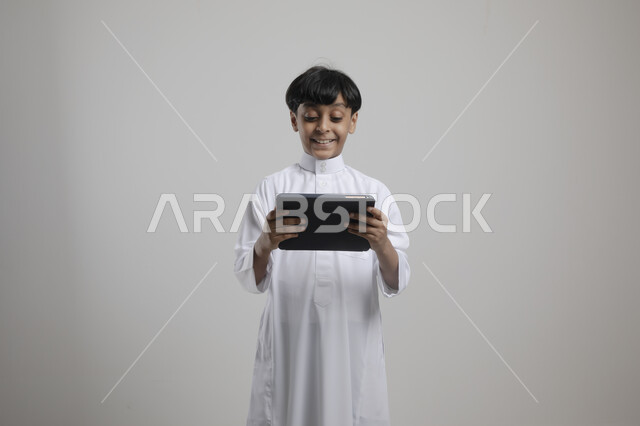 Using modern technologies to provide generations with useful information, enjoying watching favorite entertainment programs with gestures of pleasure, portrait of a smiling Saudi Arabian Gulf boy wearing a traditional dress holding a tablet in his hand, gray background