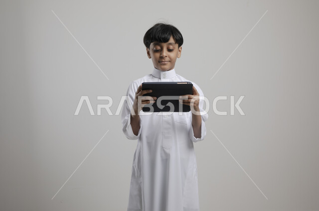 الاستمتاع في متابعه البرامج الترفيهيه المفضلة بإيماءات السرور، بورتريه لولد عربي خليجي سعودي مبتسم يرتدي الثوب التقليدي يحمل بيده جهاز التابليت، استخدام التقنيات الحديثة في تزويد الأجيال بالمعلومات المفيدة،  خلفية رمادية