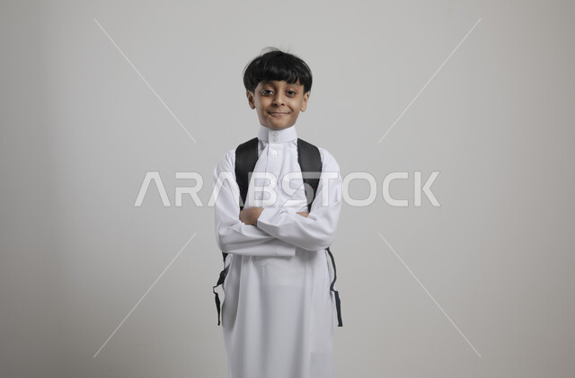 Returning to school, standing straight and looking at the camera, portrait of a Saudi Gulf Arab student wearing traditional clothing and carrying a school bag, preparing for the new school year, education in Saudi Arabia, gray background