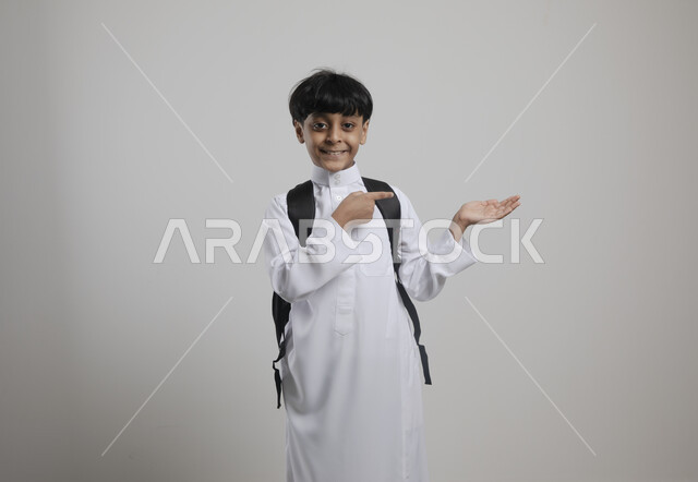 الإشارة بإصبعه السبابة إلى شيء ما ، الوقوف باستقامة والنظر إلى الكاميرا  , بورتريه لطالب عربي خليجي سعودي مبتسم يرتدي الزي التقليدي و يحمل الحقيبة المدرسية، الإستعداد للعام الدراسي الجديد، التعليم  في السعودية، العودة إلى المدارس، خلفية رمادية