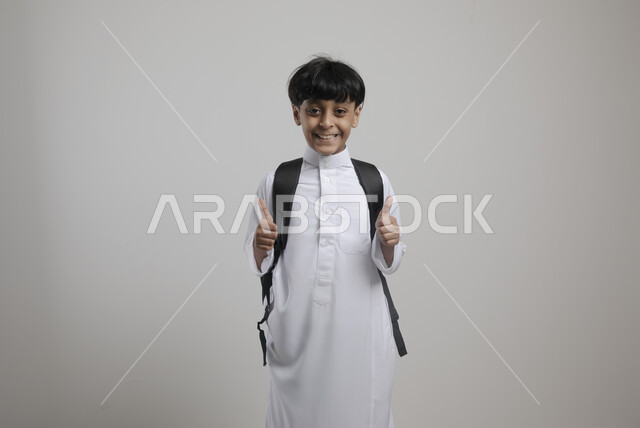 الوقوف باستعداد والنظر إلى الكاميرا  , بورتريه لطالب عربي خليجي سعودي مبتسم يرتدي الزي التقليدي و يحمل الحقيبة المدرسية، يشير بإصبعه الإبهام لأعلى للشعور بالإعجاب،  الإستعداد للعام الدراسي الجديد، التعليم  في السعودية، العودة إلى المدارس، خلفية رمادية