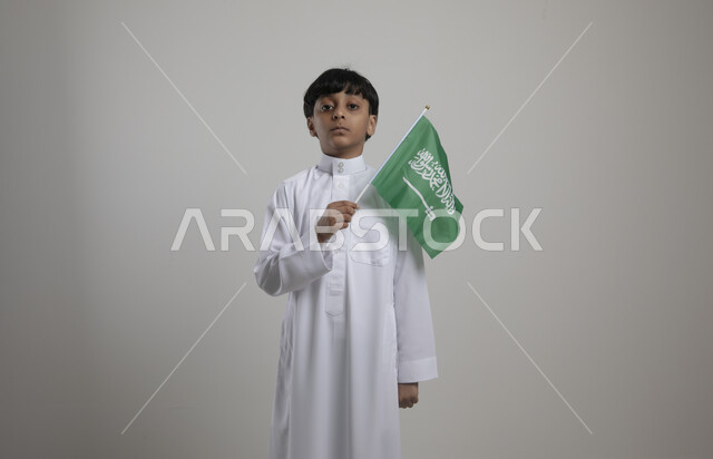 Standing straight and looking at the camera, commemorating National Day in Saudi schools, September 23, portrait of a smiling Saudi Gulf Arab child wearing traditional clothing and holding the Kingdom’s flag in his hand, Saudi Flag Day, March 11, gray background