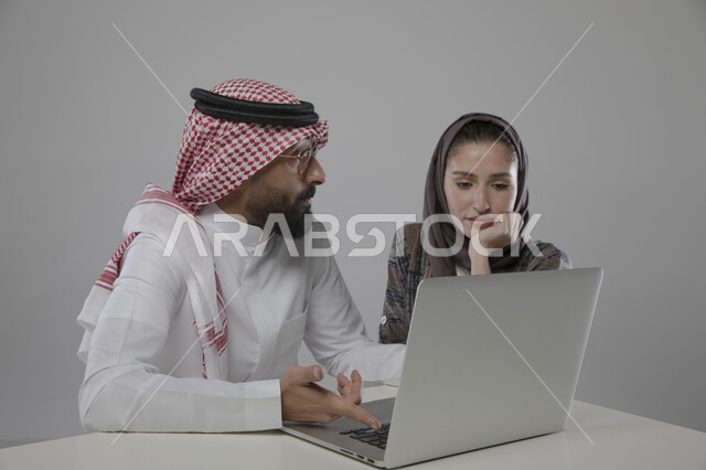 تدريب الموظفين الجدد على نظام العمل , بورتريه مقرب لزميلان عربيان سعوديان خليجيان في مقر الشركة،  مناقشة المهام  باستخدام جهاز الحاسوب المحمول ، التعاون و التفاهم بين الموظفين,   ورش واجتماعات عمل ,مهن و وظائف سعودية مكتبية , خلفية رمادية .