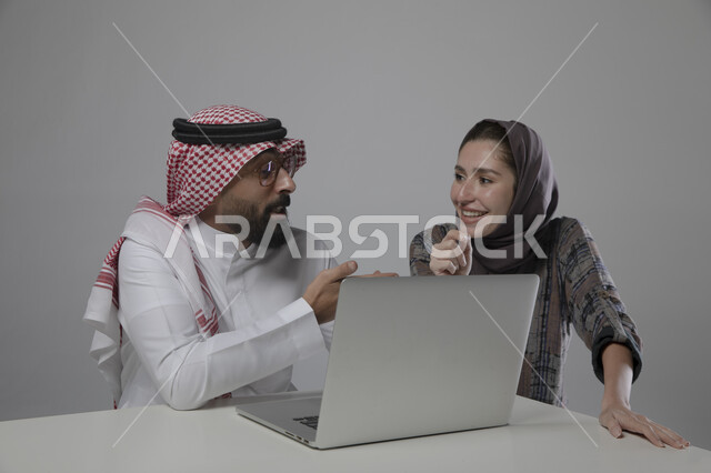 التعليم والتدريب على آلية العمل , إيماءات الرضا والسرور , بورتريه مقرب لزميلان عربيان سعوديان خليجيان في مقر الشركة،  مناقشة المهام  باستخدام جهاز الحاسوب المحمول ، التعاون و التفاهم بين الموظفين,   ورش واجتماعات عمل ,مهن و وظائف سعودية مكتبية , خلفية رمادية .