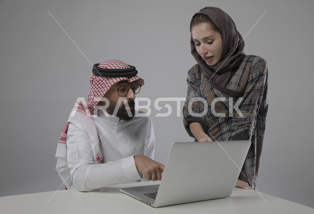 التعاون و التفاهم بين الموظفين,  ورش واجتماعات عمل ,بورتريه مقرب لزميلان عربيان سعوديان خليجيان في مقر الشركة،  مناقشة المهام باستخدام جهاز الحاسوب المحمول ، مهن و وظائف سعودية مكتبية , خلفية رمادية .