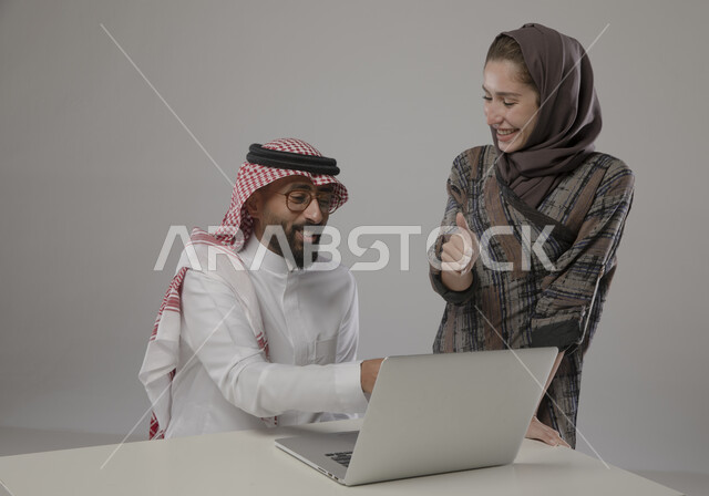 التعاون و التفاهم بين الموظفين, إيماءات السرور بزيادة الأرباح وتحقيق أهداف الشركة  , الشعور بالرضا والقبول ,  ورش واجتماعات عمل ,بورتريه مقرب لزميلان عربيان سعوديان خليجيان في مقر الشركة،  مناقشة المهام باستخدام جهاز الحاسوب المحمول ، مهن و وظائف سعودية مكتبية , خلفية رمادية .