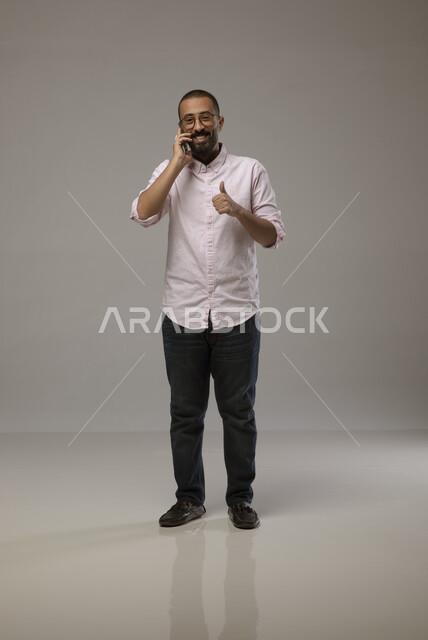 استخدام التقنيات الحديثة , إجراء مكالمات هاتفية مع الاصدقاء عبر الانترنت، بورتريه لشاب عربي خليجي سعودي مبتسم يرتدي الزي الكاجوال يدردش مع الأصدقاء باستخدام الجوال ,  رفع اصبع الابهام للدلاله على الاعجاب،  صورة الجسم بالطول الكامل، خلفية رمادية