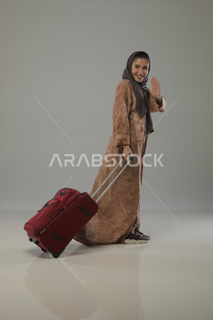 التلويح باليد وإلقاء السلام , الإستعداد لعطلة صيفية سعيدة, قضاء أوقات ممتعة في السياحة والسفر , بورتريه لسيدة عربية خليجية سعودية محجبة ترتدي عباءة وتمسك حقيبة السفر, صورة الجسم بالطول الكامل, خلفية رمادية
