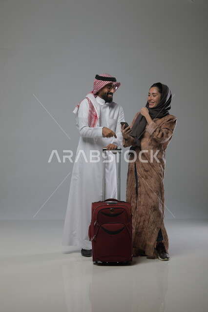 Browsing tourism and travel websites and applications via mobile phone, spending enjoyable times in tourist destinations, portrait of a Saudi Gulf Arab man holding a bag in his hand and standing next to his wife, preparing for a happy summer vacation, full-length body photo, gray background