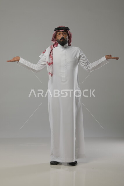 Feeling of ignorance and lack of knowledge, portrait of a Saudi Gulf Arab man wearing traditional clothing and raising his palms open upwards, standing straight and looking at the camera, marveling at something, full-length body portrait, gray background