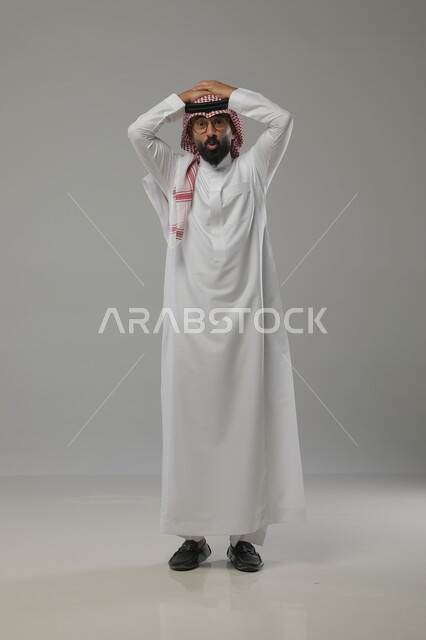 الصدمة والمفاجأة بشيء ما  , بورتريه لرجل عربي خليجي سعودي يرتدي الثوب والشماغ التقليدي و يضع يديه فوق رأسه ، النظر إلى الكاميرا والشعور بالذهول، صورة الجسم بالطول الكامل ، خلفية رمادية